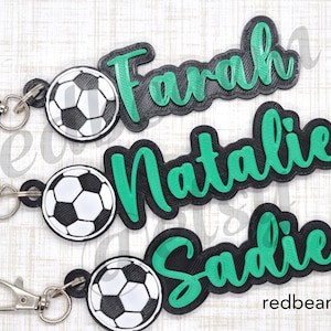 May include: Three personalized soccer-themed keychains with black and green lettering. The names "Farah", "Natalie", and "Sadie" are featured on the keychains. Each keychain has a black and white soccer ball charm.