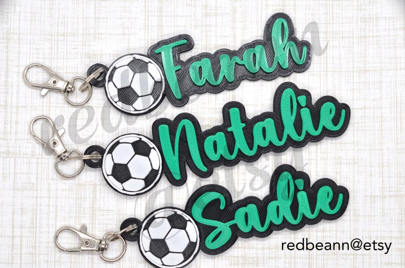 May include: Three personalized soccer-themed keychains with black and green lettering. The names "Farah", "Natalie", and "Sadie" are featured on the keychains. Each keychain has a black and white soccer ball charm.