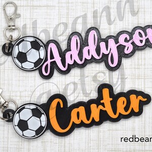 May include: Two black and white soccer ball keychains with personalized names. The keychain on the left has a pink name "Addyson" and the keychain on the right has an orange name "Carter".