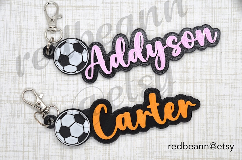 May include: Two black and white soccer ball keychains with personalized names. The keychain on the left has a pink name "Addyson" and the keychain on the right has an orange name "Carter".