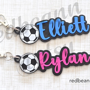 May include: Two personalized keychains with a soccer ball charm. The top keychain has a blue name tag with the name "Elliott" and the bottom keychain has a pink name tag with the name "Rylan".