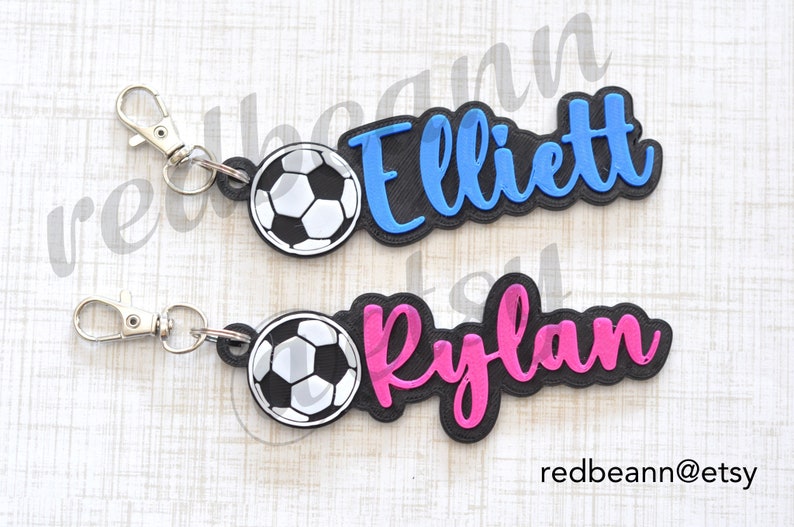 May include: Two personalized keychains with a soccer ball charm. The top keychain has a blue name tag with the name "Elliott" and the bottom keychain has a pink name tag with the name "Rylan".
