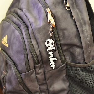 May include: A dark blue backpack with a zipper pull featuring a soccer ball and the name "Timber".