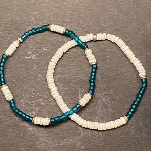 May include: Two beaded bracelets, one with teal blue beads and white shell beads, and the other with white shell beads only.