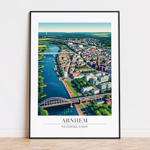 Op de afbeelding: Een ingelijste print van Arnhem, Nederland, met een gedetailleerd luchtfoto van de stad. Het kunstwerk toont een rivier, bruggen, gebouwen en groene ruimtes, met de naam van de stad en het land onderaan.