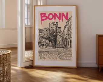 May include: A framed art print featuring a black and white sketch of a street scene in Bonn, Germany, with the city name in pink dripping letters at the top. The artwork depicts buildings, a tree, and people.