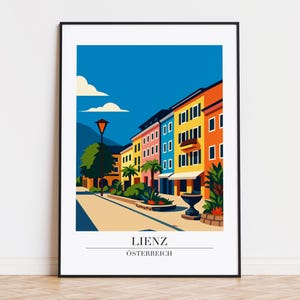 May include: A framed art print featuring a colorful illustration of Lienz, Austria. The artwork depicts a street scene with vibrant buildings in shades of orange, yellow, blue, and pink. The text "LIENZ Österreich" is displayed at the bottom.
