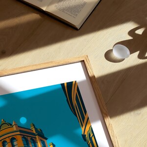 May include: A framed art print featuring a stylized architectural illustration in shades of orange, yellow, and teal. An open book and a white egg are on a wooden surface, with sunlight casting shadows.