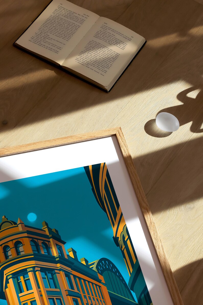 May include: A framed art print featuring a stylized architectural illustration in shades of orange, yellow, and teal. An open book and a white egg are on a wooden surface, with sunlight casting shadows.