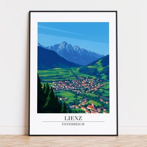 May include: A framed art print featuring a stylized landscape of Lienz, Austria. The artwork depicts a valley town nestled between green hills and towering blue mountains under a clear sky. The text "LIENZ Österreich" is at the bottom.