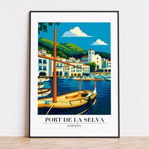 May include: A framed art print of Port de la Selva, Spain. The image features a harbor scene with a sailboat, white buildings, and green hills under a blue sky with clouds. Text at the bottom reads "PORT DE LA SELVA ESPAÑA."