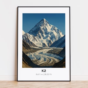 以下が含まれることがあります： K2山のフレーム入りプリント。雪に覆われた山頂が澄み切った青空の下に描かれています。画像には山の名前と標高（8,611 m）が含まれています。アートワークは黒いフレームで額装されています。