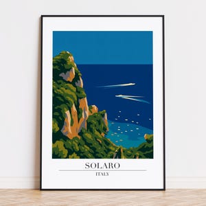 Op de afbeelding: Een ingelijste kunstdruk met een kustlandschap met een diepblauwe zee en lucht. Groene kliffen en twee witte boten zijn zichtbaar. De print bevat de tekst "SOLARO ITALY".