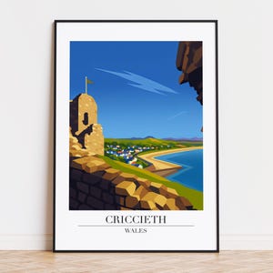 May include: A framed print of Criccieth, Wales, showing a coastal scene. The artwork features a stone castle ruin overlooking a bay with a sandy beach and a village. The sky is blue, and the print is framed in black.