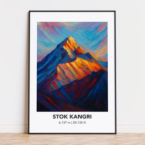 May include: A framed art print featuring a vibrant mountain landscape painting. The artwork depicts Stok Kangri, with a peak height of 20,135 ft. The mountain is painted in shades of blue, orange, and red, set against a colorful sky.