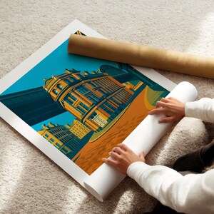 May include: A colorful art print featuring a building with a bridge in the background. The print is on a white surface, with a rolled-up print and a brown tube beside it. The print has a teal sky and orange and yellow buildings.
