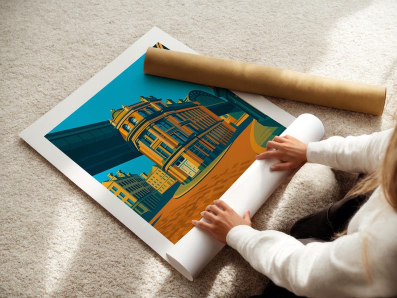 May include: A colorful art print featuring a building with a bridge in the background. The print is on a white surface, with a rolled-up print and a brown tube beside it. The print has a teal sky and orange and yellow buildings.