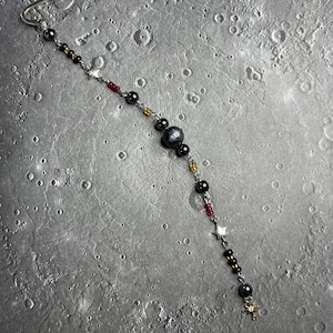 May include: A silver-toned metal bookmark featuring a dangling chain adorned with black, red, and gold beads, along with small silver star charms and a larger central dark grey bead, finished with a small gold-toned star charm at the bottom.