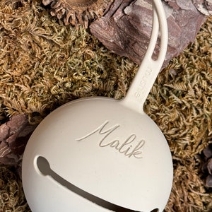 May include: A white silicone baby rattle with a loop handle. The rattle has the name "Malik" engraved on it.
