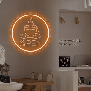 以下が含まれることがあります： コーヒーカップと「OPEN」の文字が円形に描かれたネオンサイン。サインはオレンジ色で白いアウトラインがあります。サインはカフェの壁に掛けられています。壁には「言葉が尽きたら、音楽が語る…」という文字が書かれています。