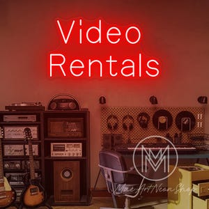 Puede incluir: Un letrero de neón rojo que dice "Video Rentals" está montado en una pared. El letrero está encima de una habitación con equipos de audio vintage, incluidas guitarras, amplificadores y un tocadiscos. La habitación tiene un ambiente de estudio de música.