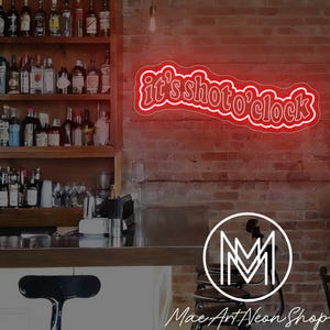 Puede incluir: Un letrero de neón rojo con el texto "it's shoto'clock" en una fuente cursiva, montado en una pared de ladrillo. El letrero está delineado en blanco. Un bar con botellas de licor es visible en el fondo.
