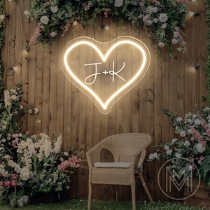 Puede incluir: Un letrero de neón blanco en forma de corazón con las iniciales "J+K" en una fuente cursiva. El letrero está colgado en una pared de madera con vegetación y flores alrededor.