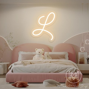 Può includere: Un'insegna al neon a forma di lettera "L" è montata su un muro sopra un letto rosa. La stanza ha una combinazione di colori tenui e pastello. Il letto è fatto con biancheria da letto bianca e ha due orsacchiotti. L'insegna è di Mac Art Neon Shop.