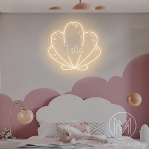 Puede incluir: Un letrero de neón con forma de concha marina con el nombre "Talia" en escritura cursiva. La luz es blanca y está montada en una pared rosa y blanca sobre una cama con ropa de cama rosa y una almohada con forma de ballena. La habitación tiene un tema decorativo.