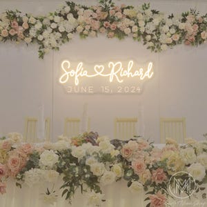 Può includere: Un tavolo da ricevimento nuziale decorato con composizioni floreali bianche e rosa. Un'insegna al neon mostra i nomi "Sofia & Richard" e la data "15 giugno 2024". Il tavolo è apparecchiato con tovaglie e sedie bianche.