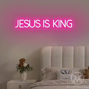 K&ouml;nnte beinhalten: Ein pinkfarbenes Neon-Schild mit dem Text "JESUS IS KING". Das Schild ist an einer rosa Wand &uuml;ber einem Bett mit wei&szlig;em Kopfteil, gestreiften Kissen und einem Nachttisch mit einer Blumenvase angebracht.