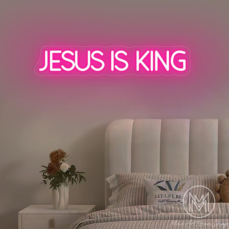 K&ouml;nnte beinhalten: Ein pinkfarbenes Neon-Schild mit dem Text "JESUS IS KING". Das Schild ist an einer rosa Wand &uuml;ber einem Bett mit wei&szlig;em Kopfteil, gestreiften Kissen und einem Nachttisch mit einer Blumenvase angebracht.