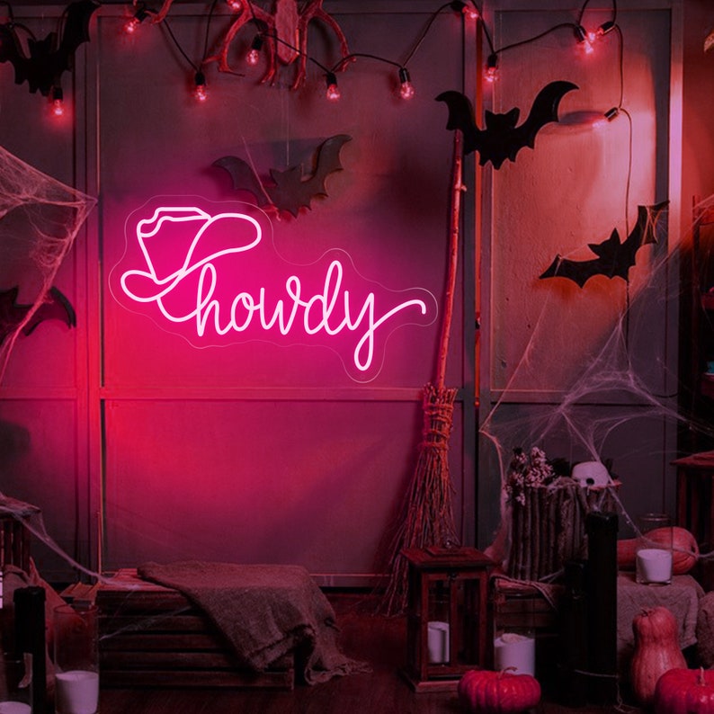 May include: Pink neon sign with a cowboy hat and the word "Howdy" in a cursive font. The sign is hanging on a wall with a dark background and a cobweb in the corner.