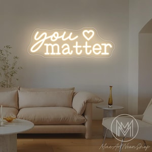 May include: A neon sign in a living room setting. The sign, in a cursive font, reads "you matter" with a heart symbol. The sign is illuminated in a warm white color. The room features a beige sofa, a side table, and a chair.