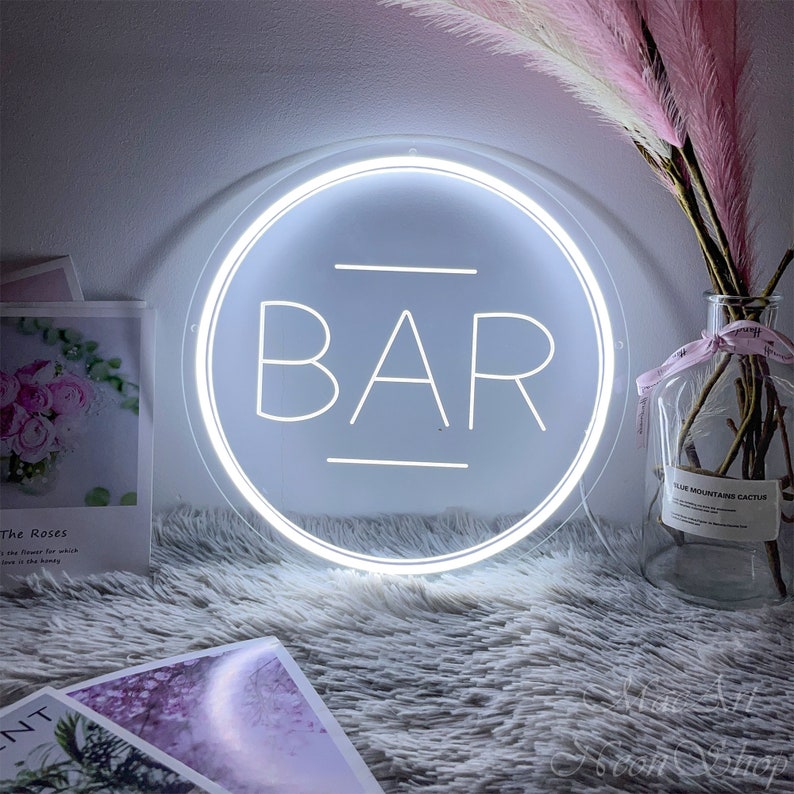 Op de afbeelding: Een wit neon-bord met het woord "BAR" in een cirkel. Het bord staat op een wit, pluche oppervlak.