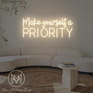 Könnte beinhalten: Ein Neonschild mit den Worten "Make yourself a PRIORITY" in kursiver Schrift. Das Schild ist an einer weißen Wand über einem weißen Sofa angebracht. Ein kleiner, hellfarbiger Tisch und ein runder Teppich befinden sich vor dem Sofa.