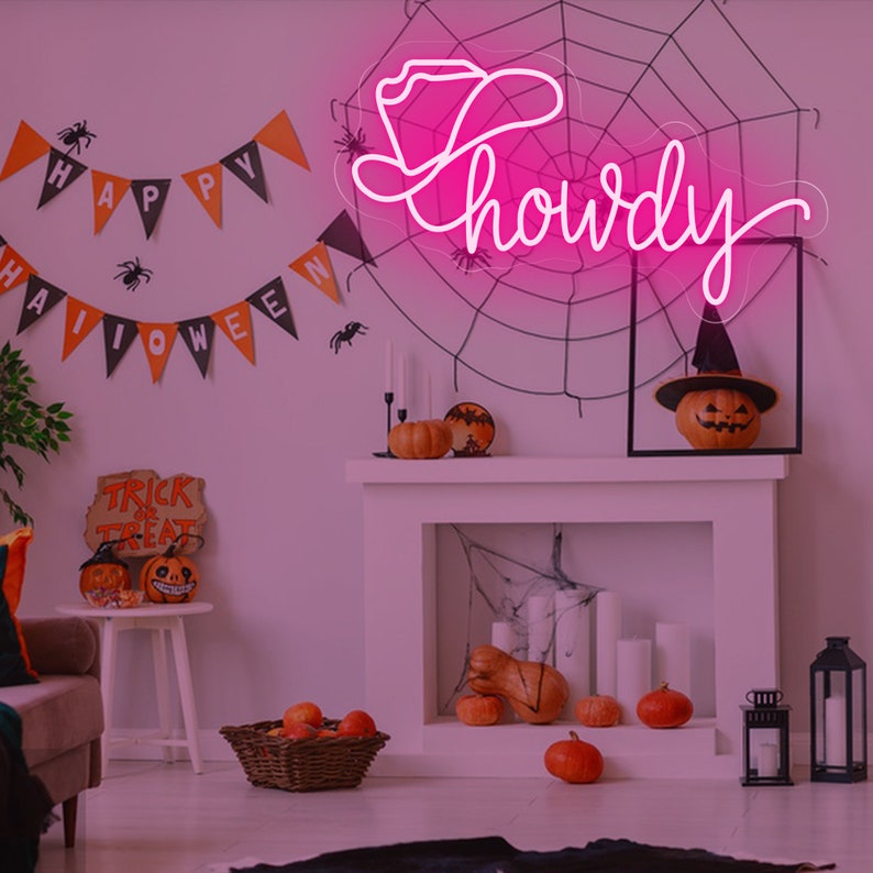 May include: Pink neon sign with the word "howdy" and a cowboy hat. The sign is hanging on a wall with a spiderweb and Halloween decorations.