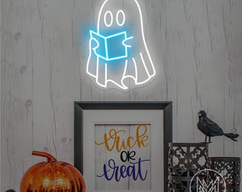 Op de afbeelding: Een witte neongeest die een blauw boek leest, gemonteerd op een houten muur. Daaronder staat op een ingelijst bord "Trick or Treat" in gouden en blauwe letters. Een decoratieve pompoen en zwarte kraaifiguren maken de Halloween-scène compleet.
