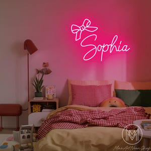Op de afbeelding: Een felroze neonbord in de vorm van een strik met de naam "Sophia" in cursief schrift. Het bord is bevestigd aan een roze muur boven een bed met rood-witte geruite beddengoed. Een rode vloerlamp en een kleine houten nachtkastje zijn ook zichtbaar.