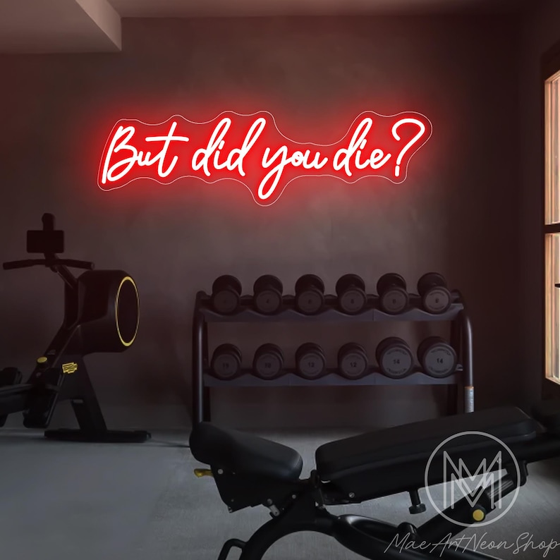Op de afbeelding: Een rood neon-bord met de tekst "But did you die?" hangt aan een muur in een sportschool. Het bord hangt boven een halterrek met zwarte halters en een zwarte halterbank.