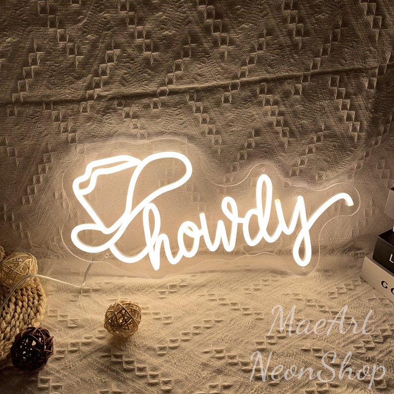 May include: White neon sign with the word "Howdy" in a cursive font, with a cowboy hat graphic to the left of the word. The sign is on a light brown textured surface.