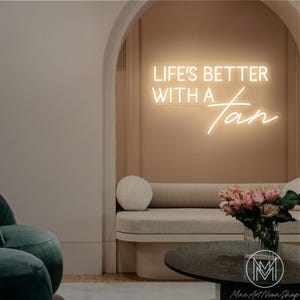 May include: A neon sign displaying the phrase "LIFE'S BETTER WITH A Tan" in a contemporary interior. The sign glows with a warm white light against a beige wall. The room includes a curved sofa, a circular coffee table, and a vase of pink flowers.