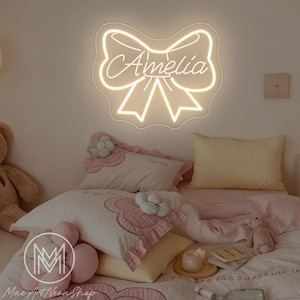 Op de afbeelding: Een neonbord in de vorm van een strik met de naam "Amelia" in cursief schrift. Het bord is aan een muur bevestigd boven een bed met roze en witte beddengoed en kussens. De kamer heeft een zachte, uitnodigende sfeer.