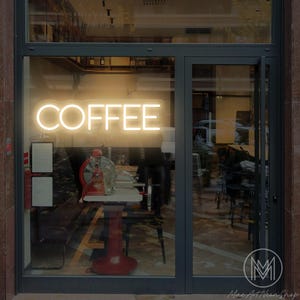 Könnte beinhalten: Ein Schaufenster zeigt ein Neon-Schild mit der Aufschrift "COFFEE" in warmem, leuchtendem Licht. Das Schild steht im Mittelpunkt, mit einem Blick ins Innere, einschließlich eines rot-basierten Tisches und einer alten Kaffeemühle.