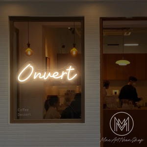 Puede incluir: Un escaparate con un letrero de neón que dice "Ouvert" en una fuente cursiva. Debajo del letrero, se ven las palabras "Coffee Dessert". El logotipo de la tienda es una "M" blanca dentro de un círculo, con "Mac Art Neon Shop" debajo.