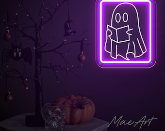 Op de afbeelding: Een paars neonbord met een witte omtrek van een geest die een boek leest. Het bord is in een vierkant frame en heeft de tekst "Mae Art Neon Shop" eronder.