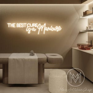 Könnte beinhalten: Ein Spa-Interieur mit einer Massageliege, einem Waschbecken und einem Neon-Schild mit der Aufschrift "THE BEST CURE is a Manicure". Der Raum ist in neutralen Tönen mit einer modernen Ästhetik eingerichtet. Ein kleiner, runder, strukturierter Hocker befindet sich im Vordergrund.