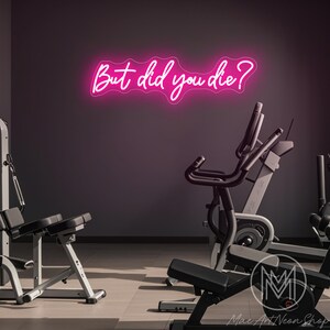 Op de afbeelding: Een roze neonbord met de tekst "But did you die?" hangt aan een muur in een sportschool. Het bord hangt boven een hometrainer en andere fitnessapparatuur.