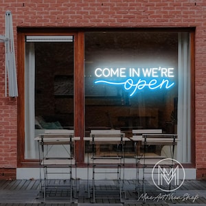 Könnte beinhalten: Ein Neonschild in einem Schaufenster zeigt die Worte "COME IN WE'RE open" in weißer und blauer Schrift. Das Schild ist in einem Holzrahmen angebracht, mit Sitzgelegenheiten im Freien darunter. Der Hintergrund ist eine Backsteinmauer.