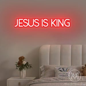 K&ouml;nnte beinhalten: Ein rotes Neonzeichen mit der Aufschrift &bdquo;JESUS IS KING&ldquo; ist &uuml;ber einem Bett an der Wand angebracht. Der Raum ist mit einem Nachttisch, Blumen und Kissen dekoriert. Das Schild erhellt den Raum mit einem warmen Schein.